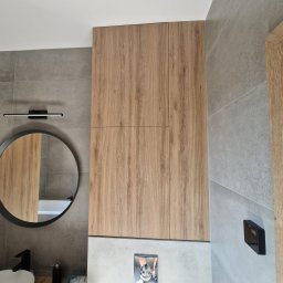 FIX WOOD ZABUDOWY MEBLOWE - Wysoka szafka łazienkowa z drewnianymi frontami, szara ściana z płytek, okrągłe lustro z czarną ramą i minimalistyczna lampa nad nim, widoczny fragment umywalki.