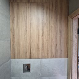FIX WOOD ZABUDOWY MEBLOWE - Trzy drewniane szafki wiszące nad szarymi płytkami w łazience, z widocznym przyciskiem spłuczki i fragmentem drewnianej futryny drzwi.