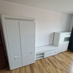 Biały zestaw mebli do salonu z szafą, szafką RTV i witryną, na drewnianej podłodze, widoczne instrukcje montażu na szufladzie.