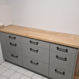 FIX WOOD ZABUDOWY MEBLOWE - Szara komoda z dziewięcioma szufladami i drewnianym blatem, czarne uchwyty, na tle białej ściany z fakturą i białej podłogi z płytek.