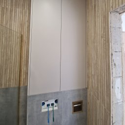 FIX WOOD ZABUDOWY MEBLOWE - Nowoczesna zabudowa toalety podtynkowej: białe szafki, drewnopodobne panele ścienne i szara glazura. Widoczne elementy instalacji przed montażem armatury.