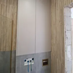 Nowoczesna zabudowa toalety podtynkowej: białe szafki, drewnopodobne panele ścienne i szara glazura. Widoczne elementy instalacji przed montażem armatury.