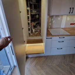 FIX WOOD ZABUDOWY MEBLOWE - Biała kuchnia z otwartą szafką spiżarnianą i podświetlanymi półkami. Widoczny fragment dłoni otwierającej drzwi szafki.