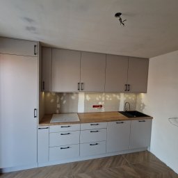 FIX WOOD ZABUDOWY MEBLOWE - Jasnoszara zabudowa kuchenna z drewnianym blatem i czarnymi uchwytami, widoczne niedokończone wykończenie ściany nad blatem, w tle surowy sufit z wystającymi kablami.