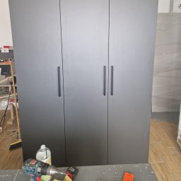 FIX WOOD ZABUDOWY MEBLOWE - Szara, trzydrzwiowa szafa z długimi, pionowymi uchwytami na tle podłogi z paneli. Na pierwszym planie wiertarka i akcesoria montażowe.