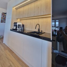 FIX WOOD ZABUDOWY MEBLOWE - Nowoczesna kuchnia z białymi szafkami i drewnianymi frontami, oświetlona listwami LED. Na blacie ekspres do kawy i czarny zlew z baterią. Widoczne odbicie osoby robiącej zdjęcie.