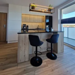 Nowoczesna kuchnia z wyspą i hokerami. Drewniane fronty szafek oświetlone LED, czarny blat i dodatki. Minimalistyczny design z roślinami w wiszącej konstrukcji.