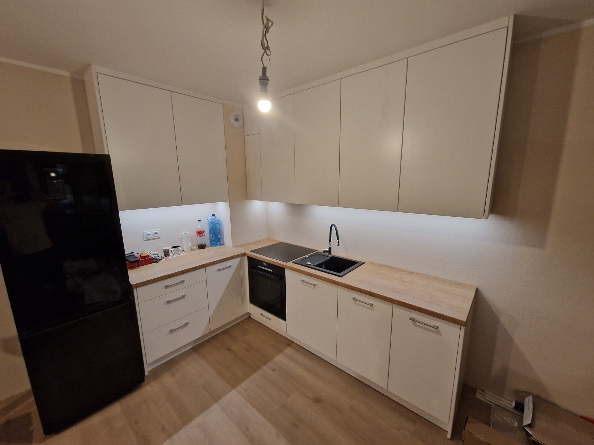 Nowoczesna, jasna kuchnia z białymi frontami szafek i drewnianym blatem, czarny zlew i płyta indukcyjna, widoczna lodówka z czarnym frontem, surowa żarówka zwisająca z sufitu.