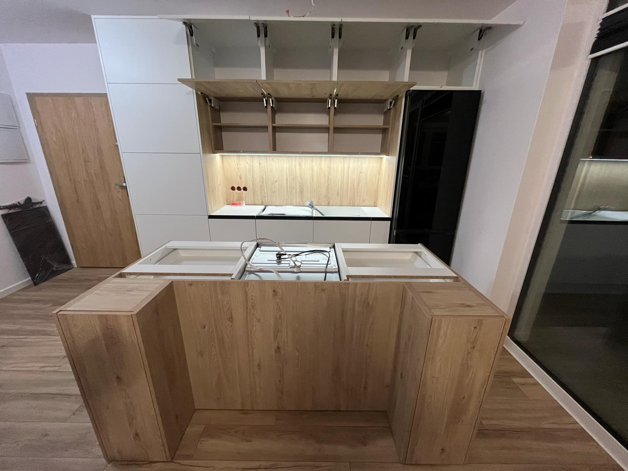 Wnętrze kuchni w trakcie montażu: wyspa kuchenna z drewnianym wykończeniem, widoczne okablowanie płyty grzewczej, białe szafki wiszące z otwartymi frontami, podświetlenie LED nad blatem.