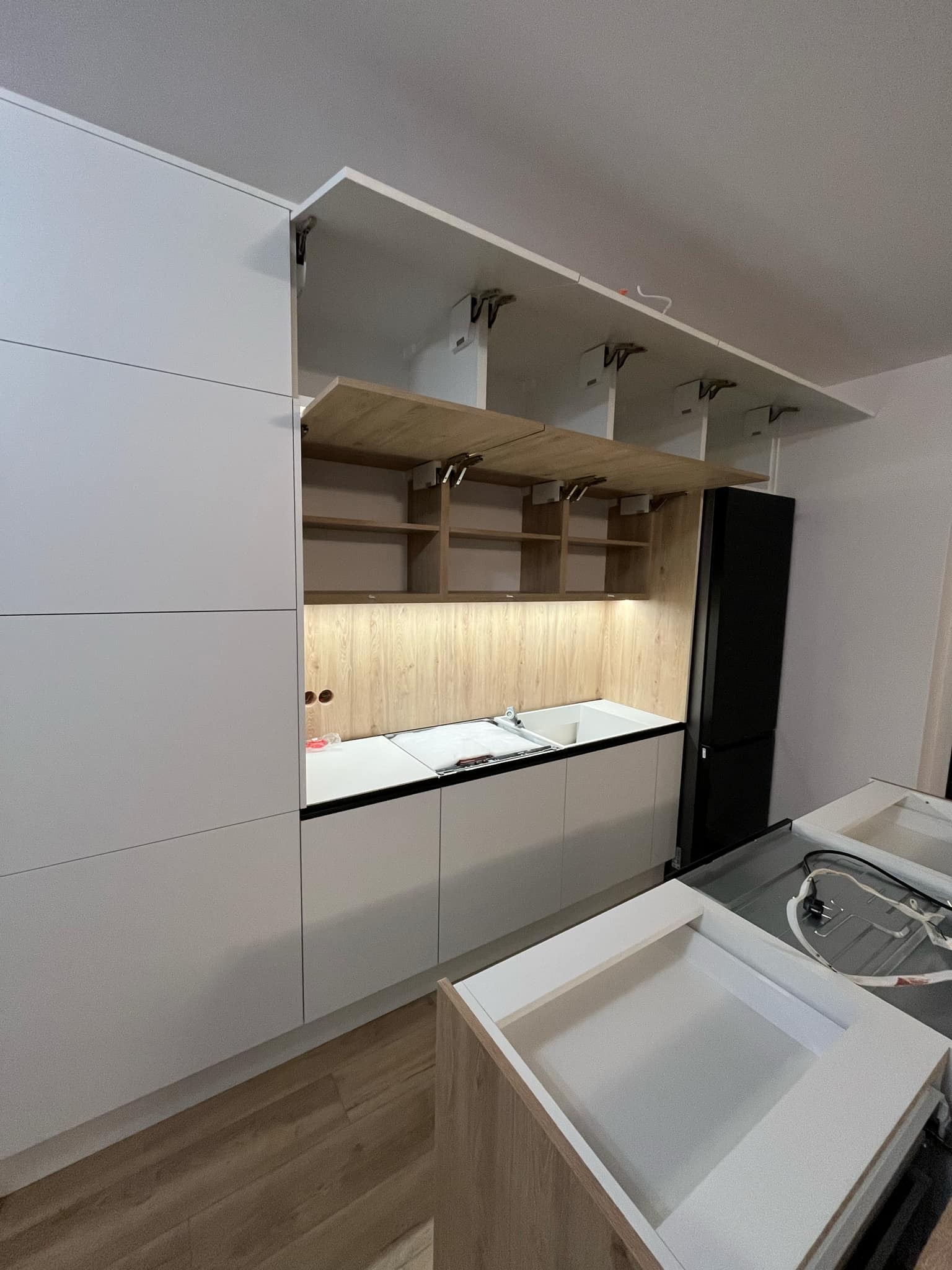 Nowoczesna kuchnia z białymi frontami, otwartymi szafkami górnymi z drewnianym wnętrzem i podświetleniem LED, w trakcie montażu, widoczne niedokończone elementy i instalacje.
