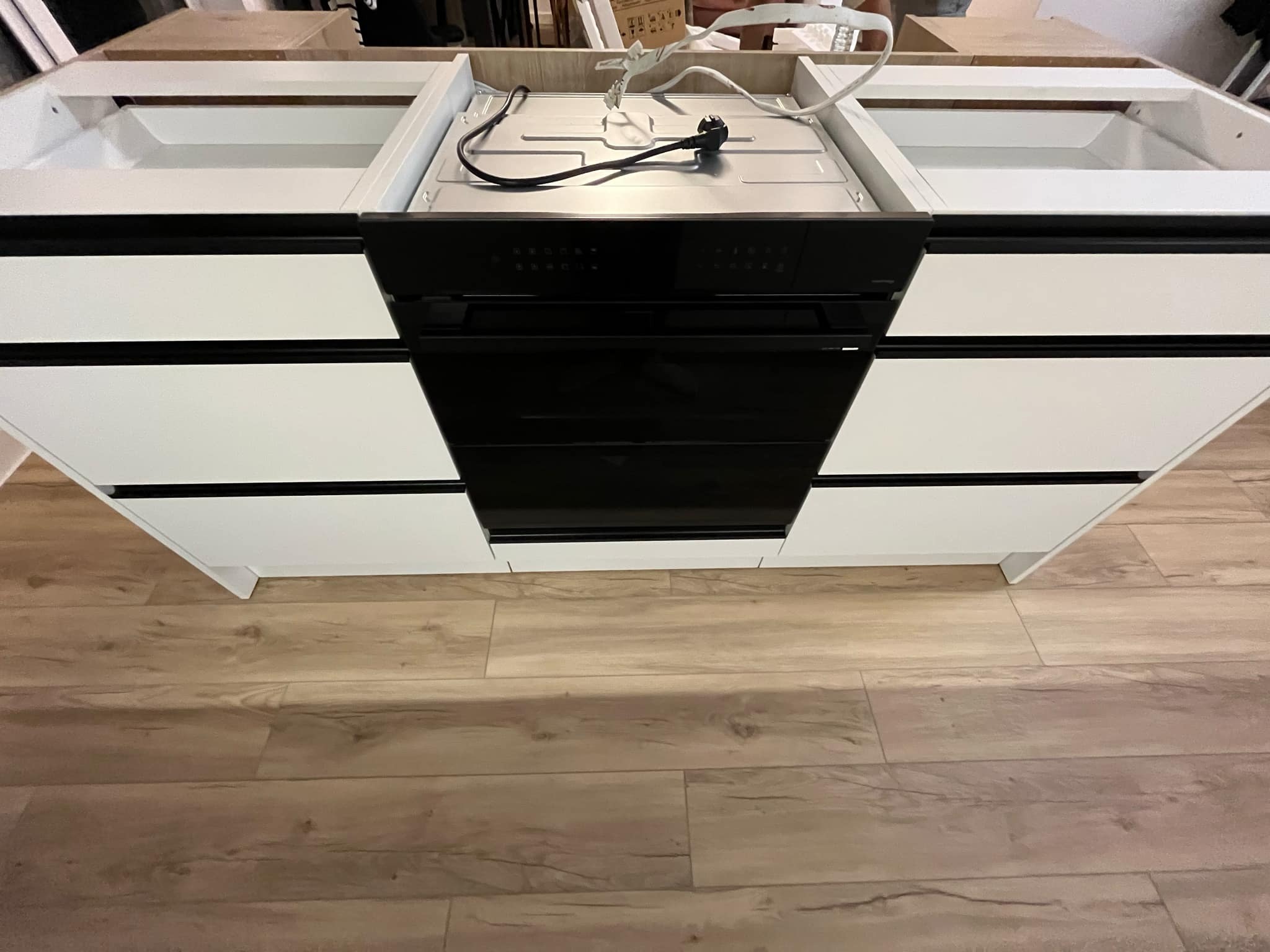 Częściowo zmontowany moduł kuchenny z białymi szafkami i czarnym piekarnikiem w trakcie instalacji, widoczne kable i elementy montażowe, podłoga wyłożona panelami.