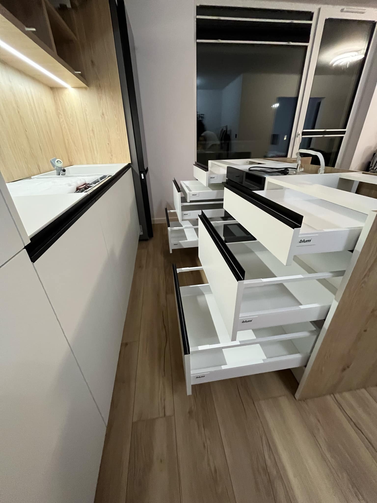 Nowoczesna kuchnia z białymi frontami i czarnymi uchwytami, otwarte szuflady z systemem Blum, widok na drewnianą podłogę i okno.