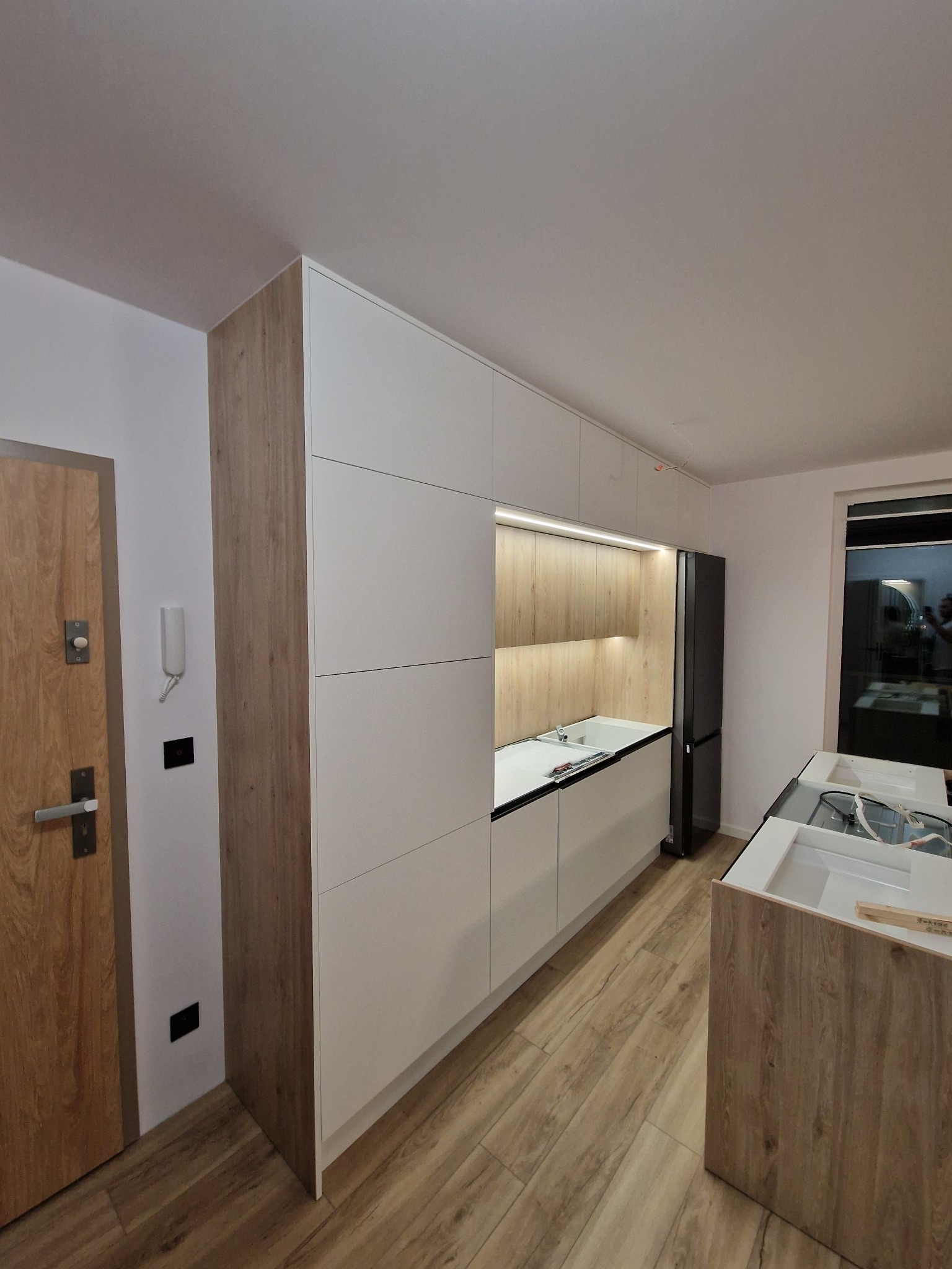 Nowoczesna kuchnia z białymi frontami i drewnianymi akcentami, zintegrowanym oświetleniem LED, widoczna lodówka i fragment wyspy kuchennej w trakcie montażu, na podłodze panele imitujące drewno.