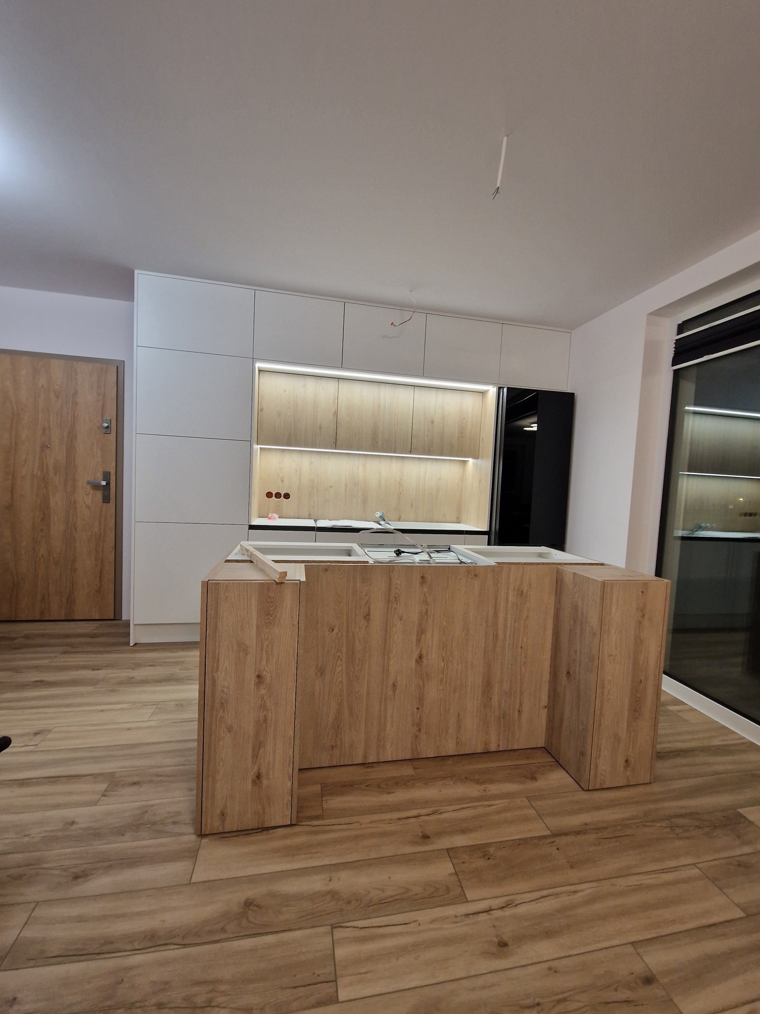 Wnętrze kuchni w trakcie montażu, widoczna wyspa kuchenna z drewnianym wykończeniem, białe szafki wiszące z oświetleniem LED, oraz fragment podłogi z paneli.