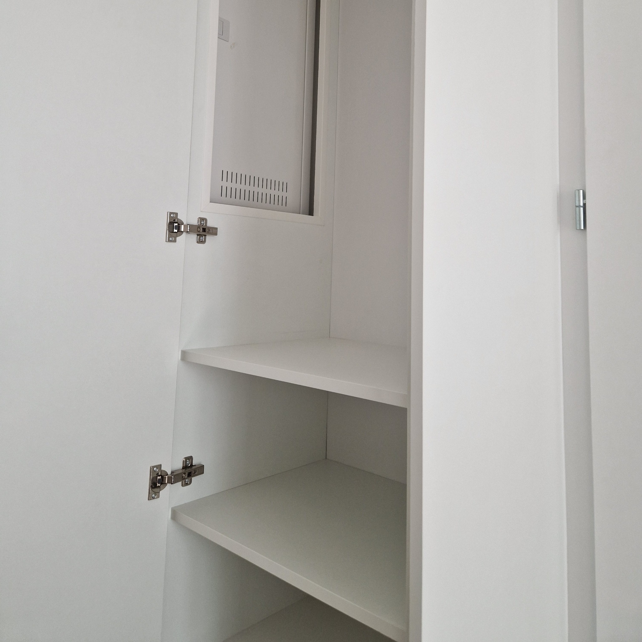 Wnętrze białej szafy wnękowej z półkami i widocznymi zawiasami. Na górze szafy, nad półkami, znajduje się otwór wentylacyjny.