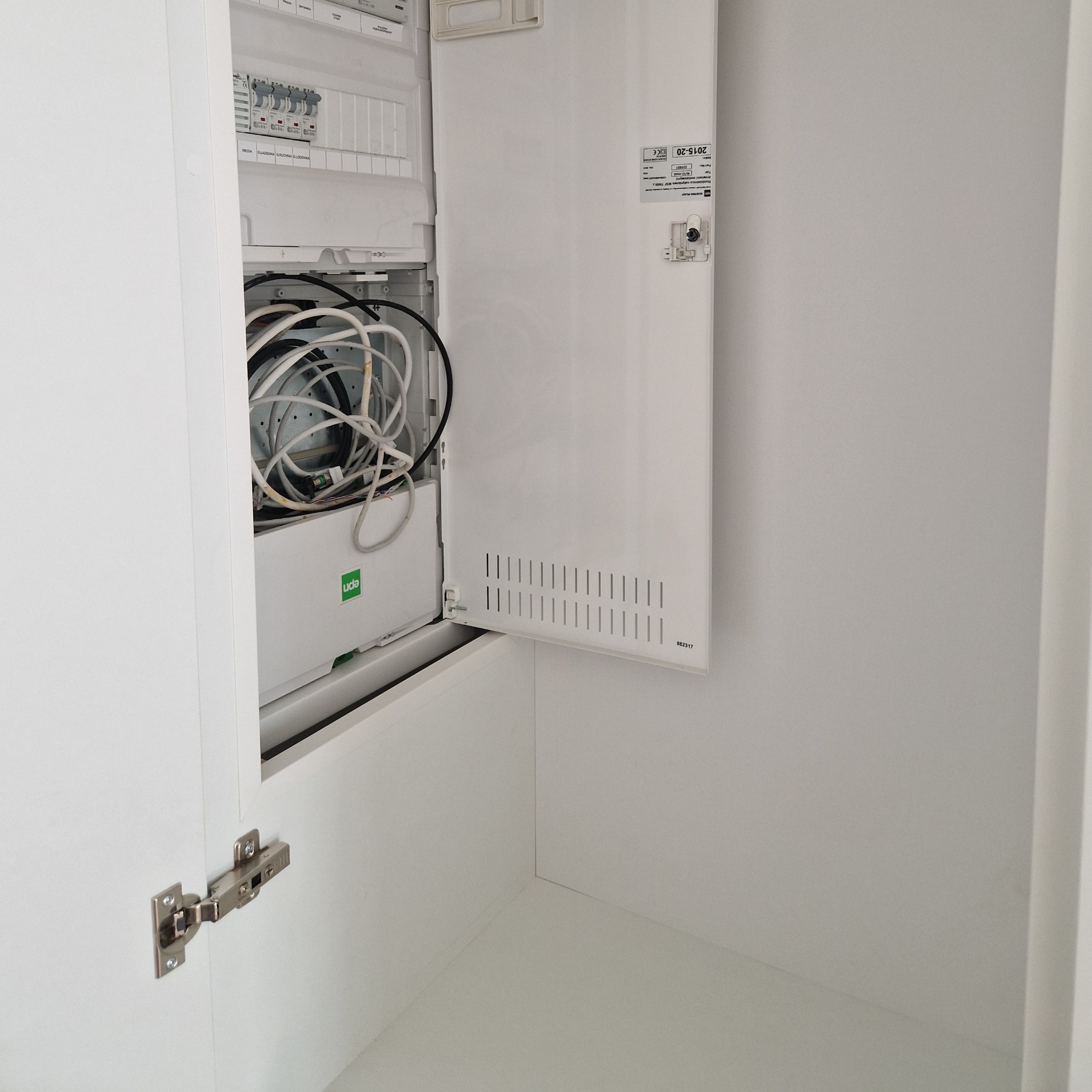 Otwarta, biała szafka z widoczną instalacją elektryczną i kablami wewnątrz, zamontowana na ścianie. Widoczny fragment zawiasu.