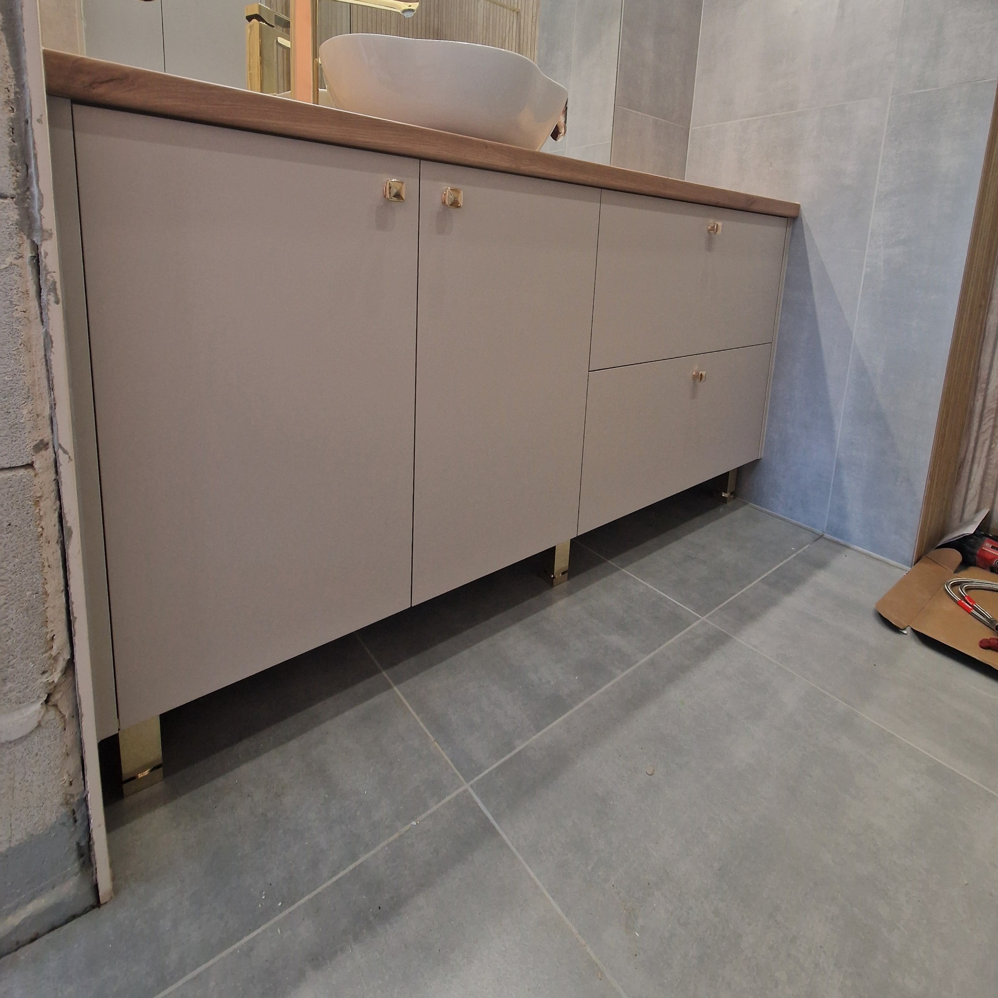 Szara szafka łazienkowa z drewnianym blatem i złotymi uchwytami, z umywalką na blacie. Widoczne narzędzia na podłodze sugerują świeży montaż.