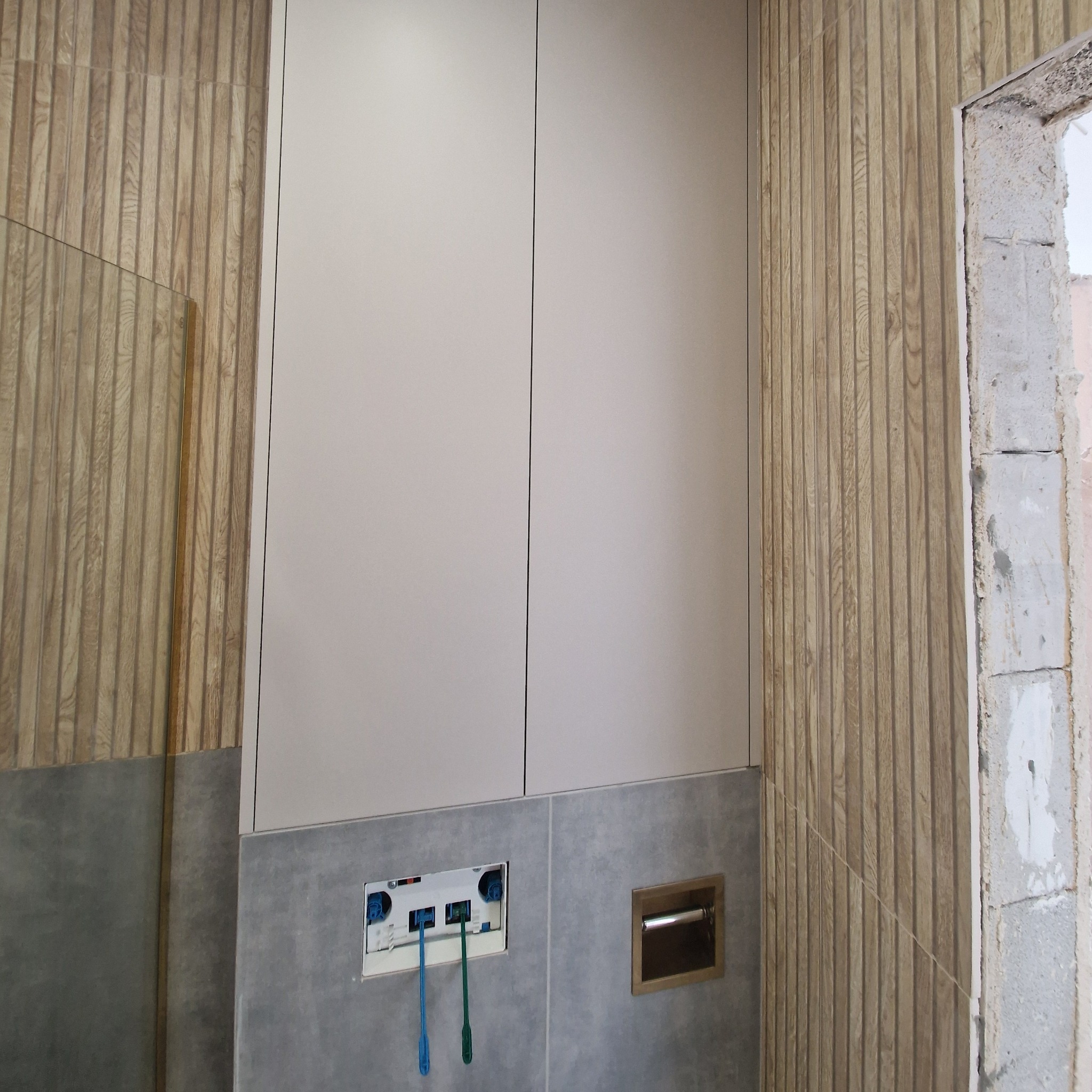 Nowoczesna zabudowa toalety podtynkowej: białe szafki, drewnopodobne panele ścienne i szara glazura. Widoczne elementy instalacji przed montażem armatury.
