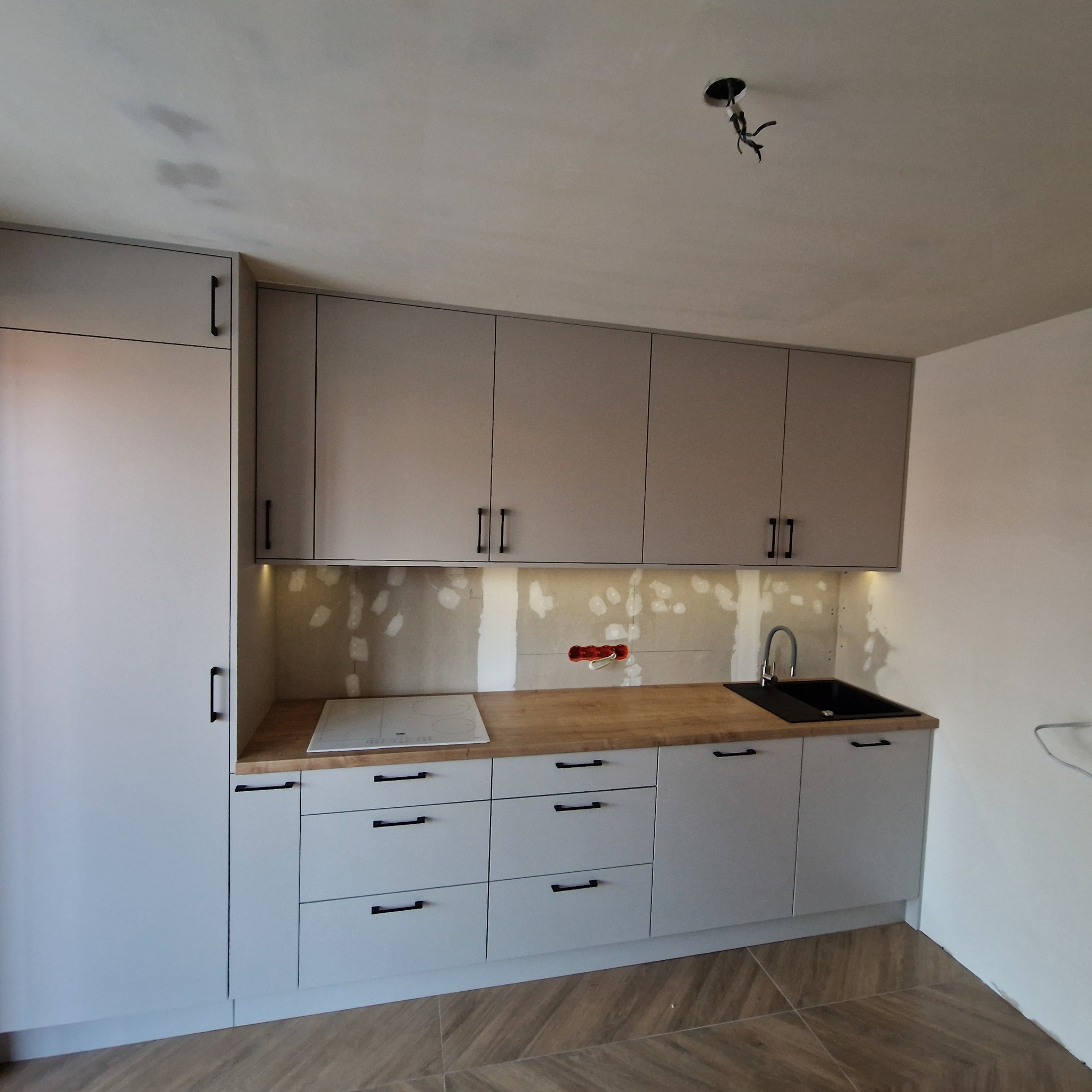 Jasnoszara zabudowa kuchenna z drewnianym blatem i czarnymi uchwytami, widoczne niedokończone wykończenie ściany nad blatem, w tle surowy sufit z wystającymi kablami.