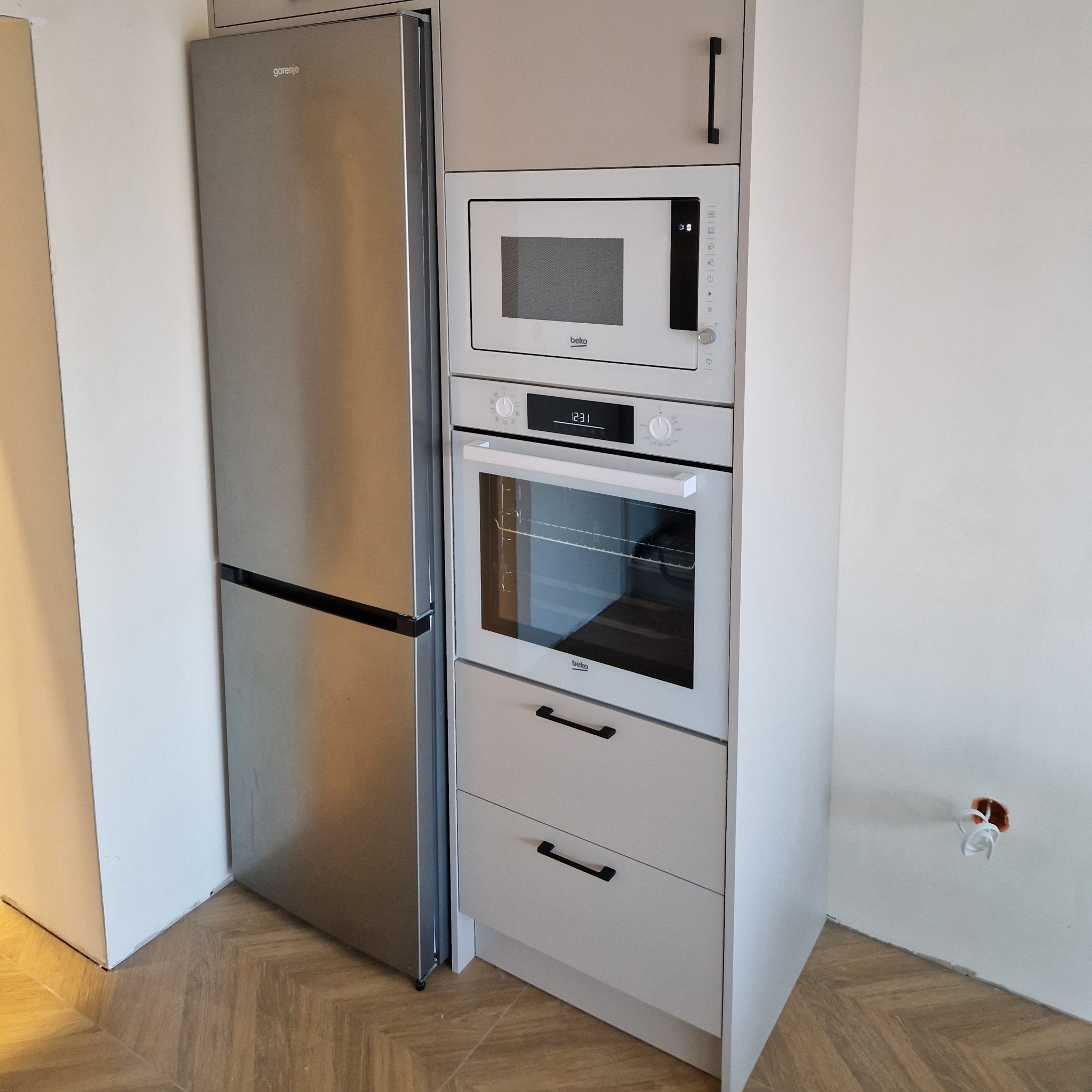 Kompaktowa zabudowa kuchenna w jasnej tonacji: lodówka Gorenje, piekarnik i mikrofalówka Beko. Na podłodze parkiet ułożony w jodełkę. Widoczne gniazdko elektryczne.