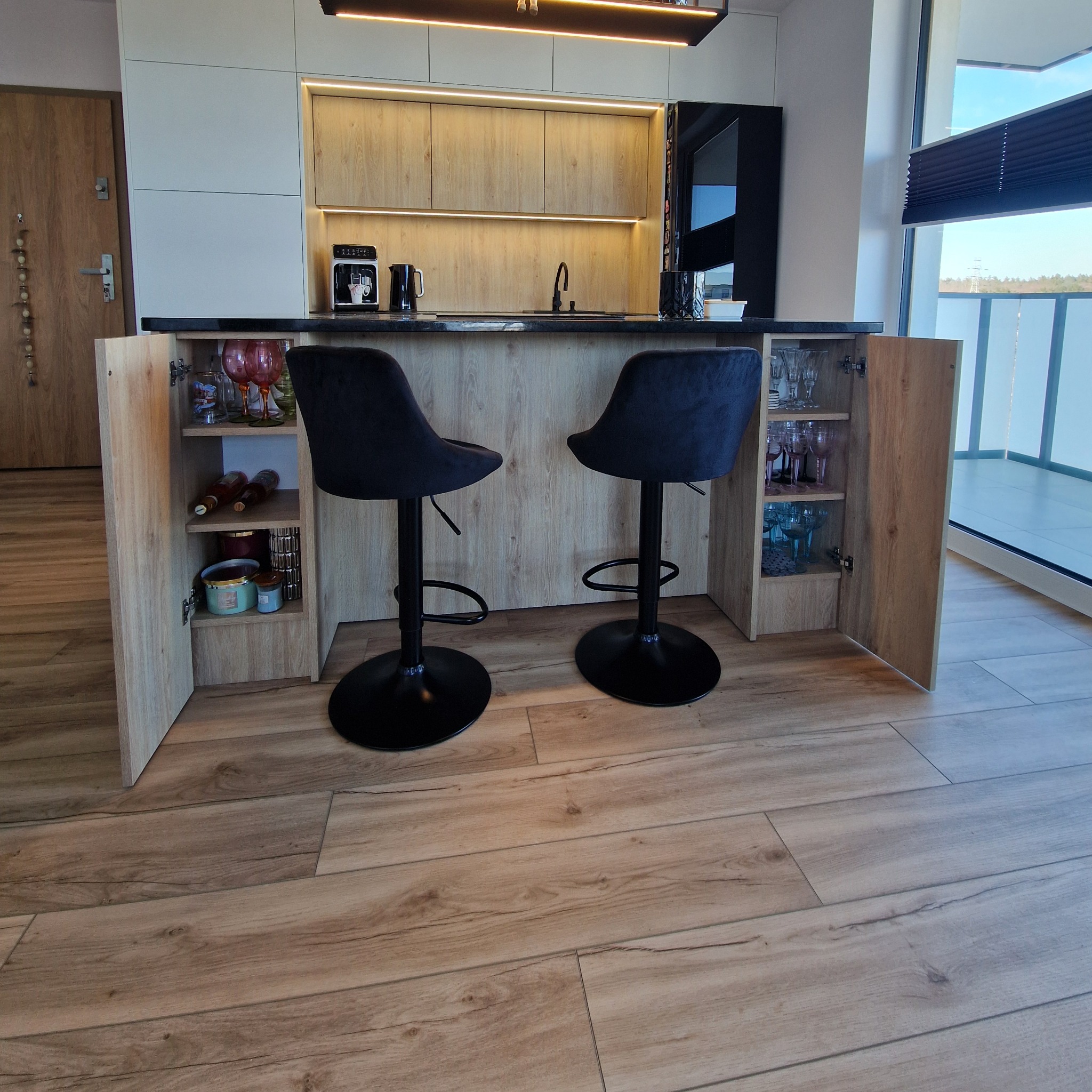 Nowoczesna kuchnia z wyspą, drewniane fronty szafek, czarne hokery. Otwarta szafka z dekoracyjnym szkłem i oświetleniem LED. Podłoga z paneli imitujących drewno.