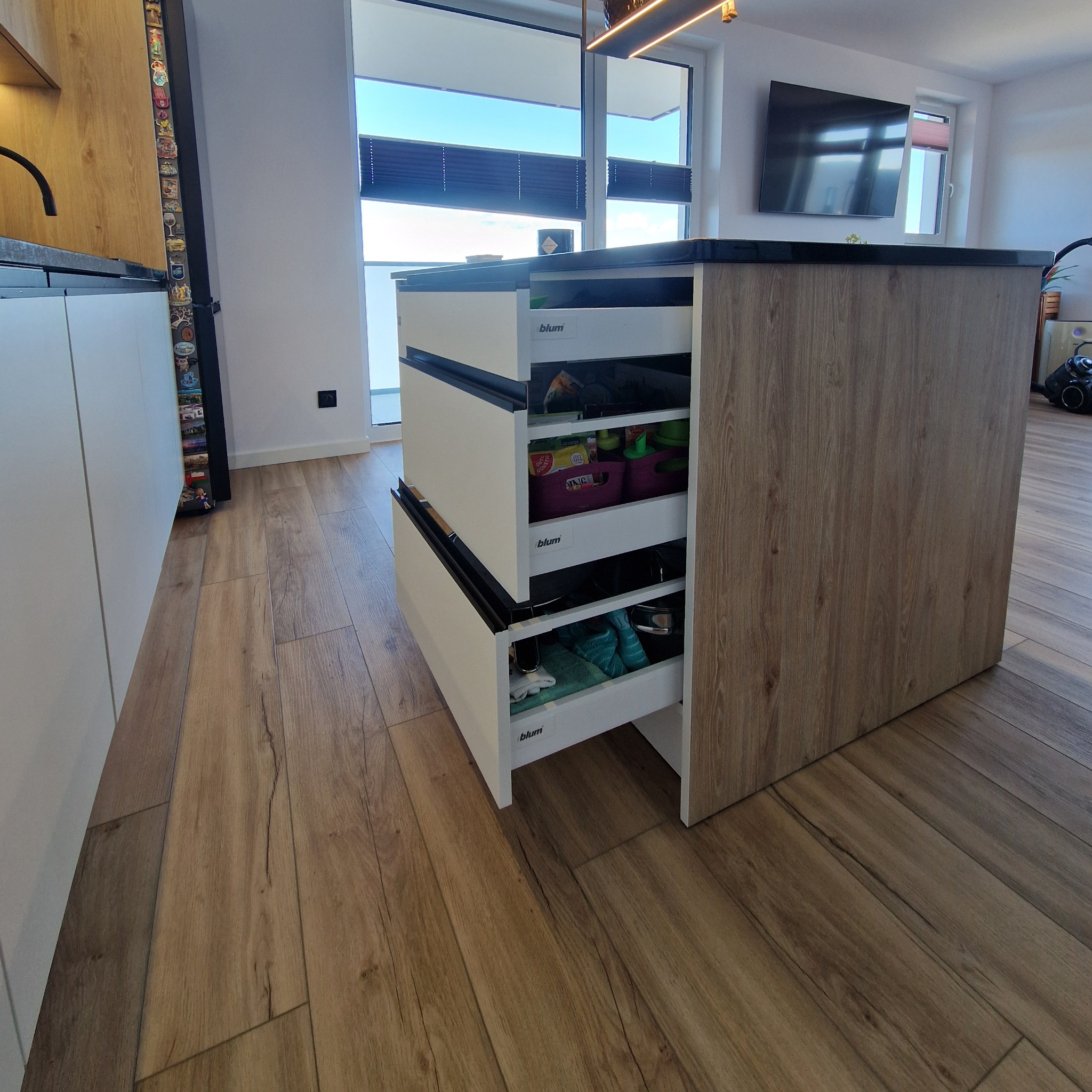 Otwarta szuflada w wyspie kuchennej z systemem Blum, ukazująca zawartość. Blat czarny, fronty białe, korpus w odcieniu drewna. Podłoga z paneli imitujących drewno.