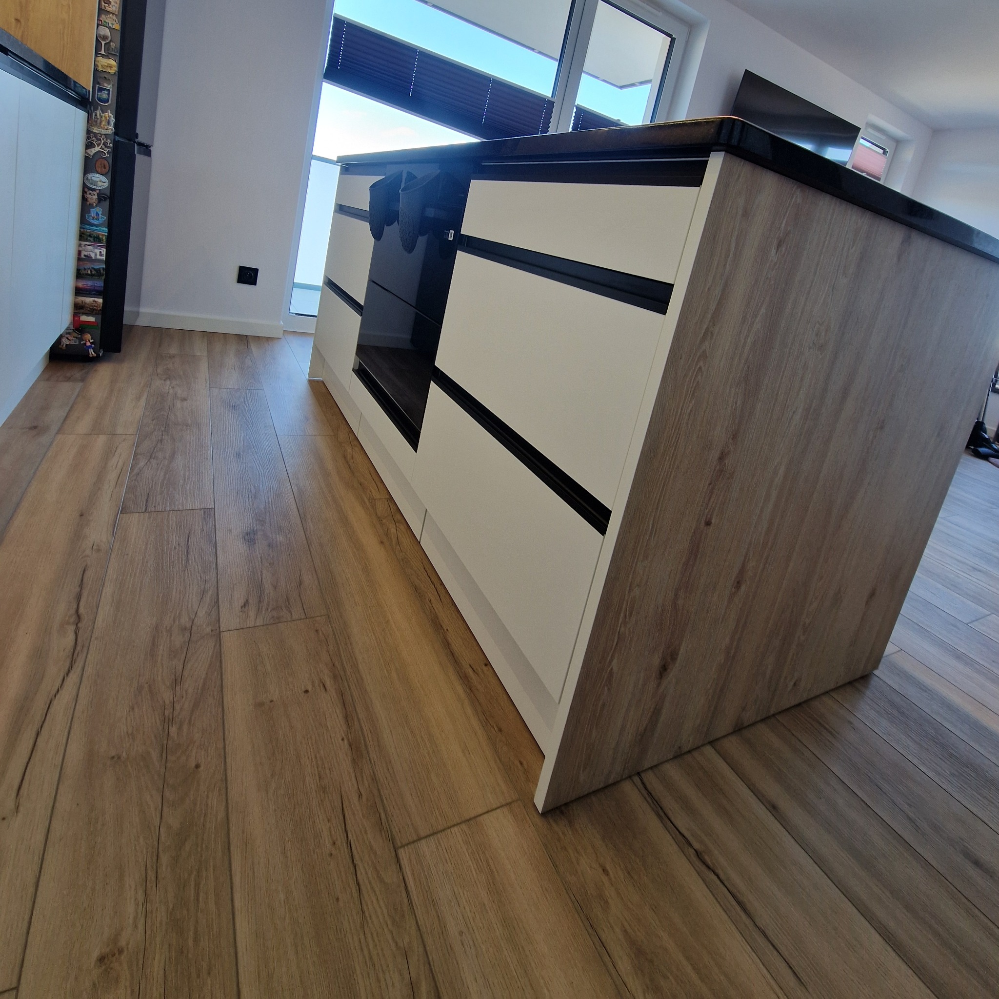 Nowoczesna wyspa kuchenna z białymi frontami i drewnopodobnym wykończeniem, czarny blat, widoczne panele podłogowe, minimalistyczny design, duże okno w tle.