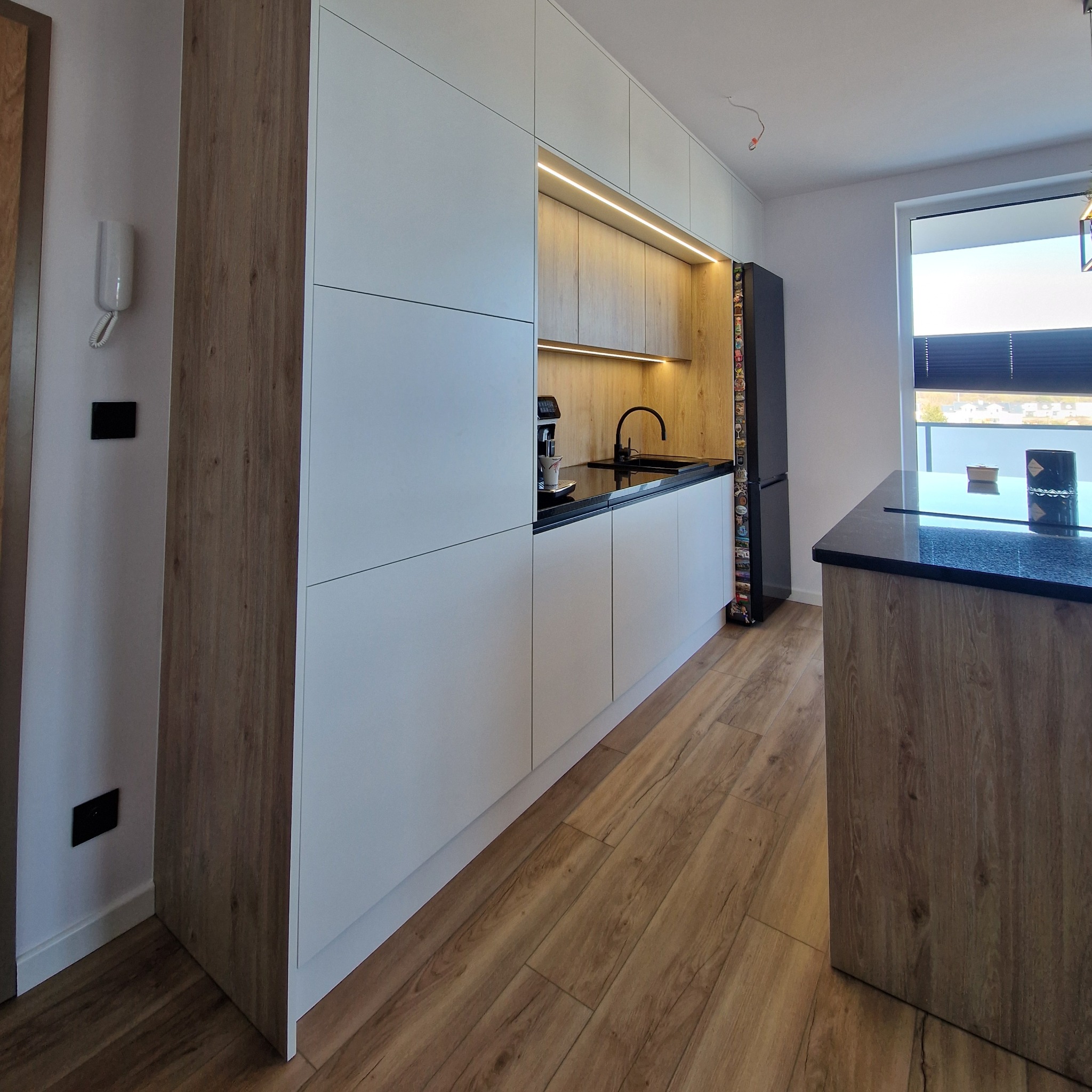 Nowoczesna kuchnia z białymi szafkami i drewnianymi akcentami, oświetleniem LED, czarnym blatem i podłogą z paneli. Widoczny fragment wyspy kuchennej.