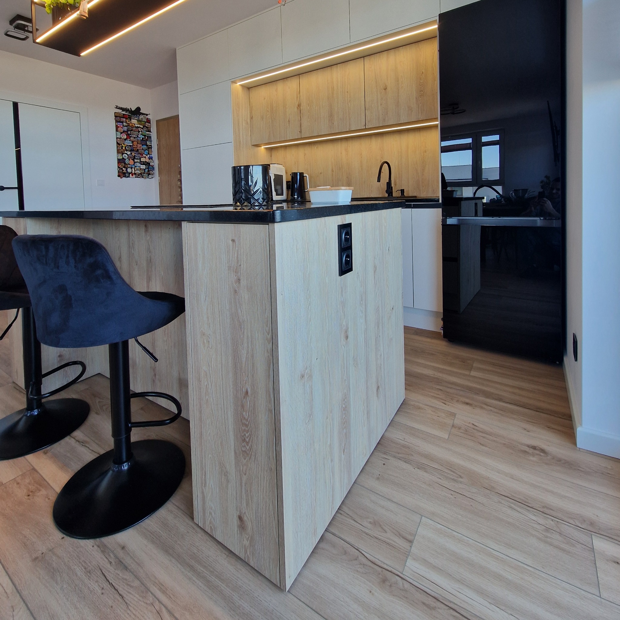 Nowoczesna kuchnia z wyspą o drewnianym wykończeniu, czarnym blatem i hokerami. Widoczne oświetlenie podszafkowe i czarna lodówka. Podłoga wyłożona panelami.