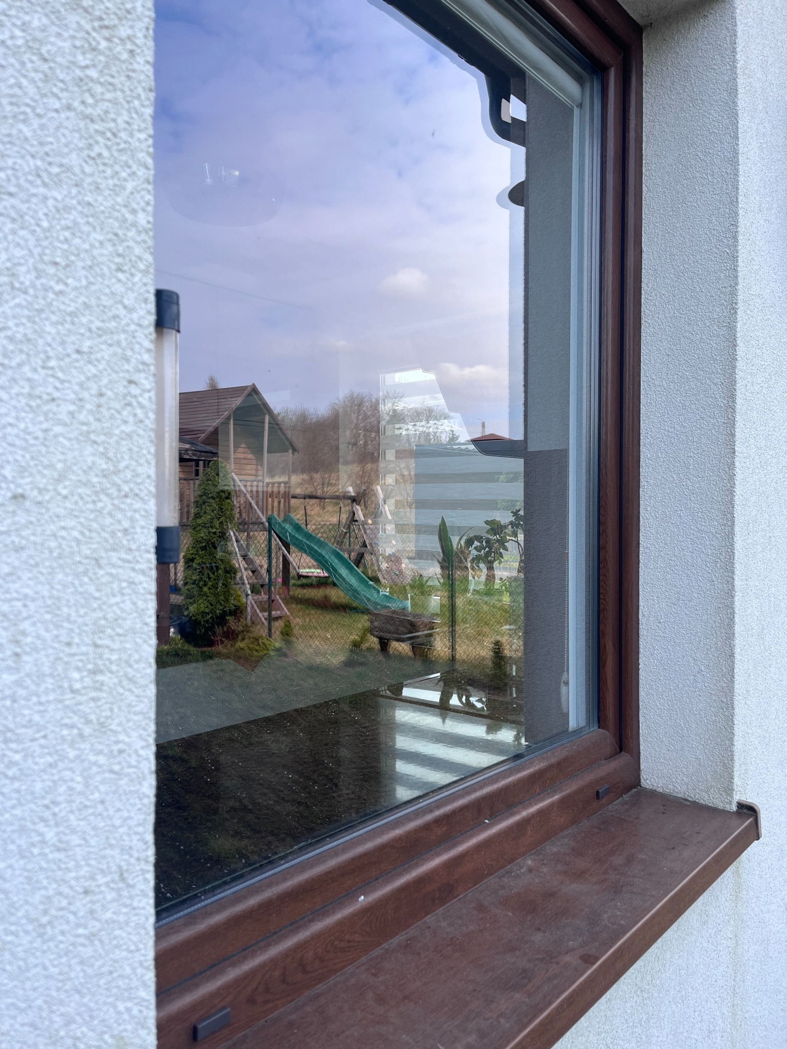 Widok z okna na plac zabaw z domkiem, zjeżdżalnią i huśtawką, odbijający się w szybie. Widoczny brązowy parapet i biała elewacja budynku.
