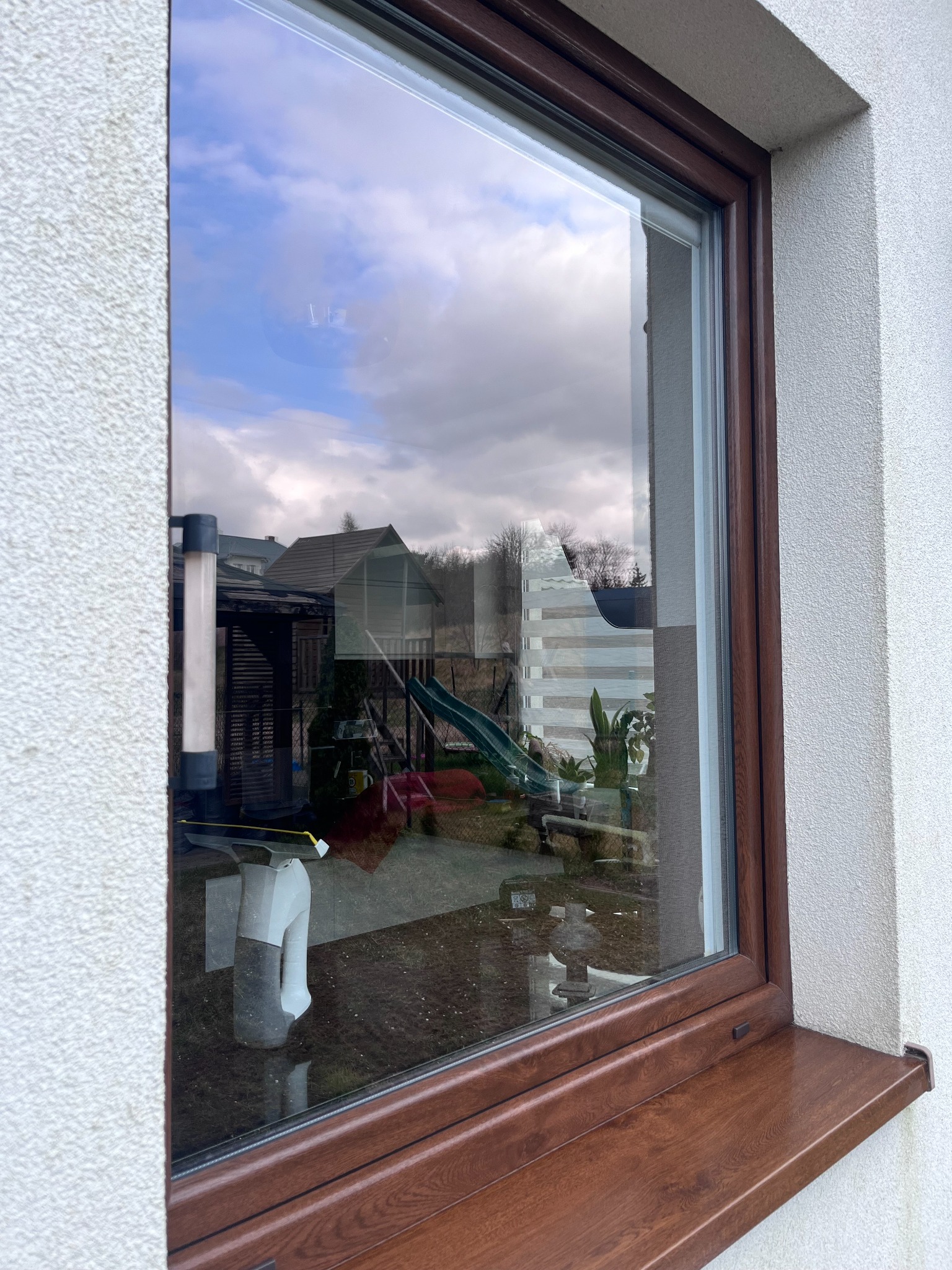 Brązowe okno w Gdańsku z widocznym odbiciem ogrodu, w rogu szyby leży biało-szary spryskiwacz do mycia okien.