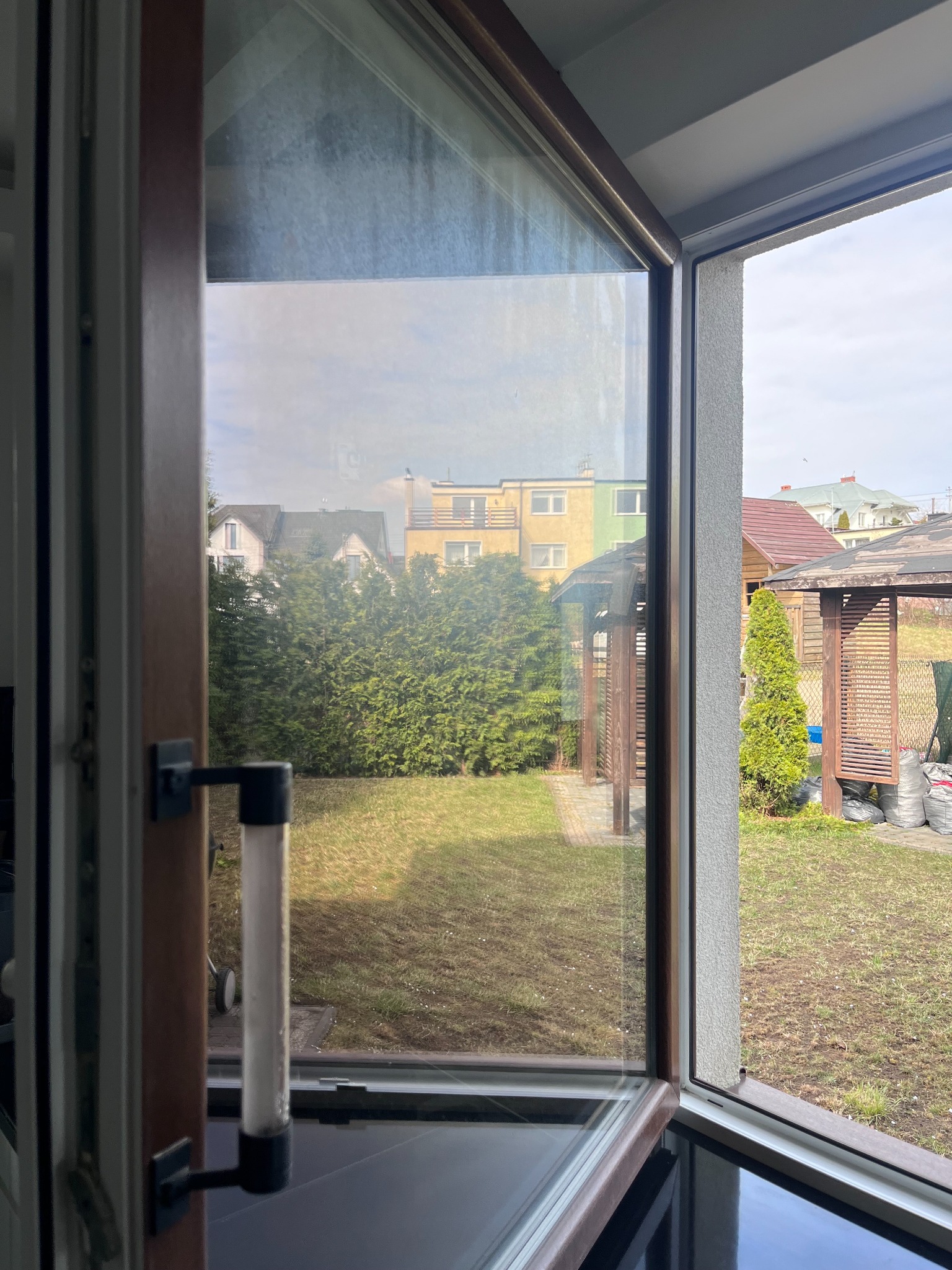 Widok przez brudną szybę okienną na ogród z tujami, altanami i domami w tle, uchylone okno z widocznym mechanizmem otwierania.