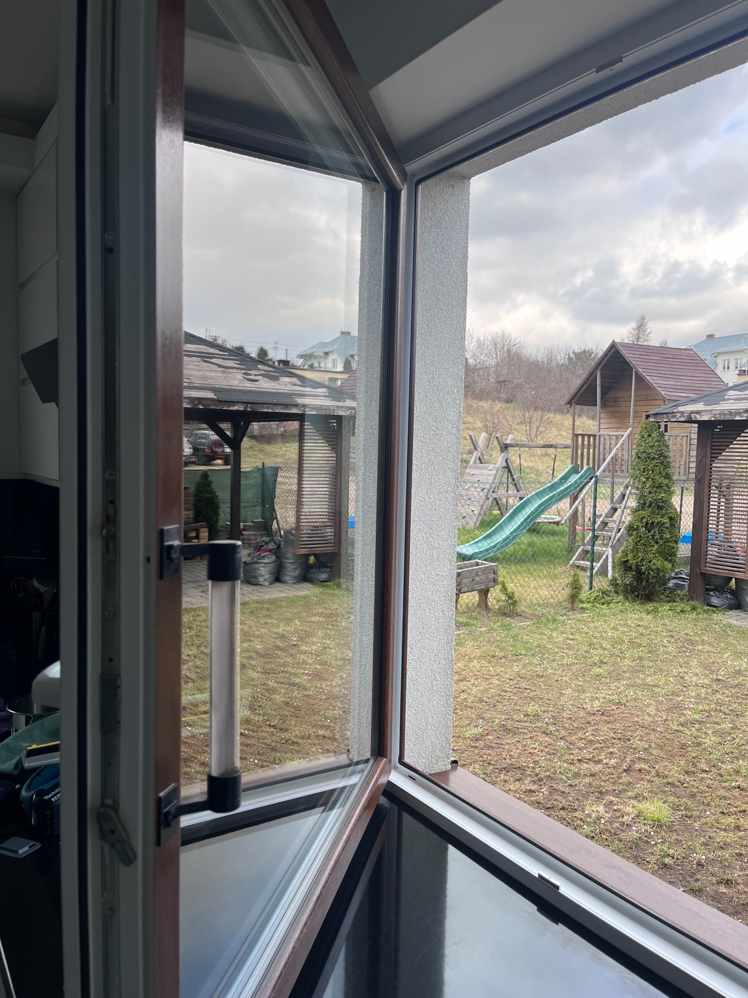 Widok z otwartego okna na podwórko z drewnianym placem zabaw i altaną, pochmurne niebo.
