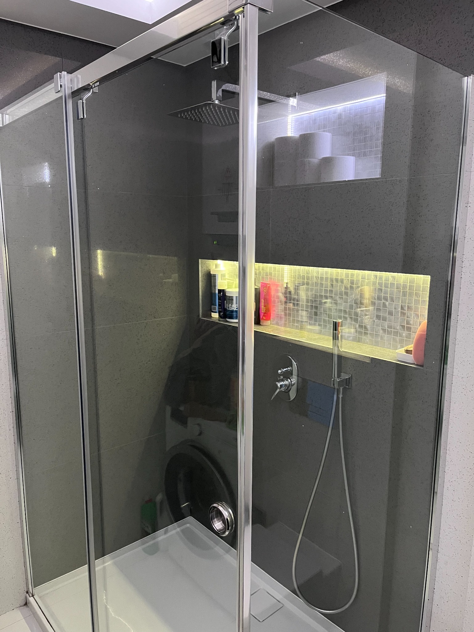 Szklana kabina prysznicowa z deszczownicą i wnękami w ścianie wyłożonymi mozaiką, podświetlonymi ledami, z widoczną pralką w tle.