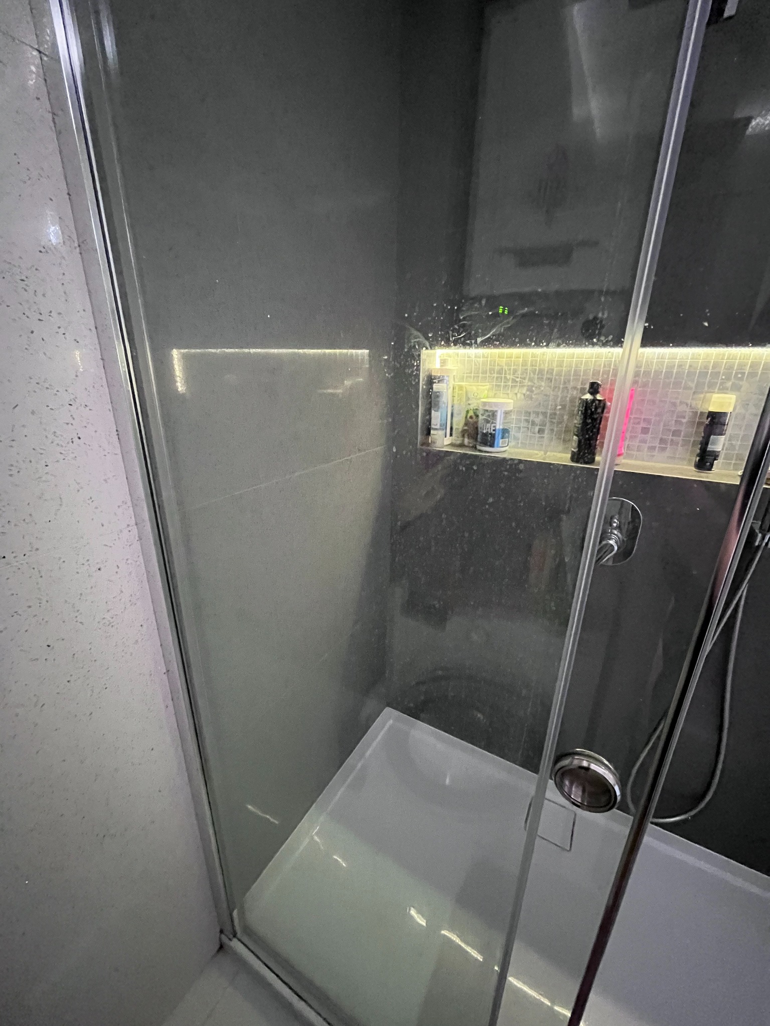 Szklana kabina prysznicowa z widocznymi smugami i zaciekami, z półką na kosmetyki podświetloną listwą LED, ukazująca potrzebę czyszczenia