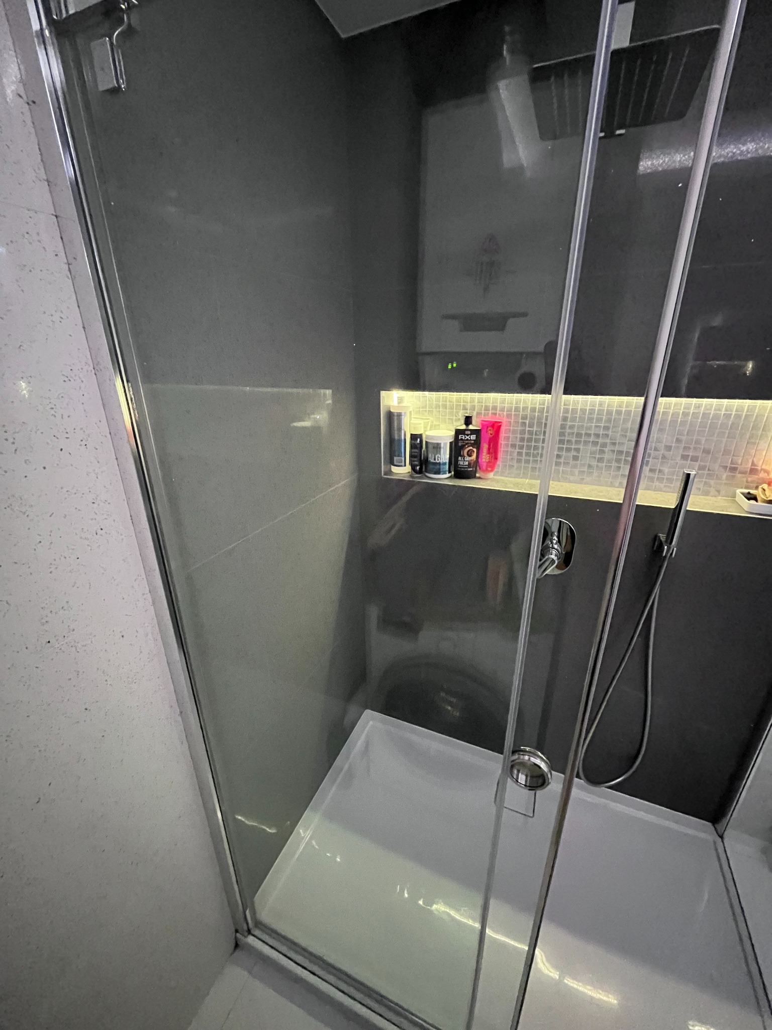 Szklana kabina prysznicowa z widoczną półką z kosmetykami, baterią i słuchawką prysznicową, a w tle pralka.