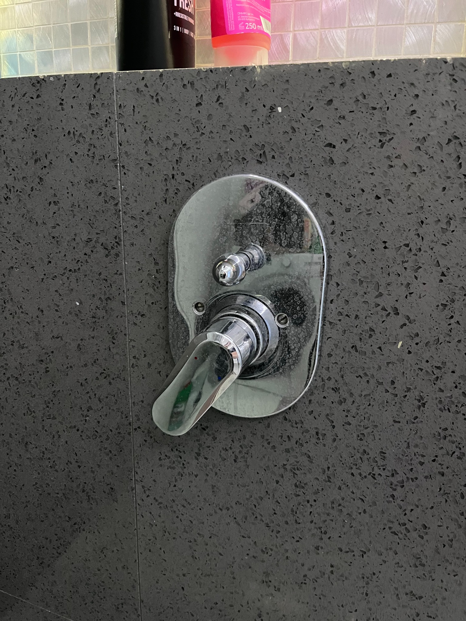 Chromowana bateria prysznicowa z dwoma pokrętłami zamontowana na ciemnej, teksturowanej ścianie prysznica, widoczne ślady po wodzie na chromie.