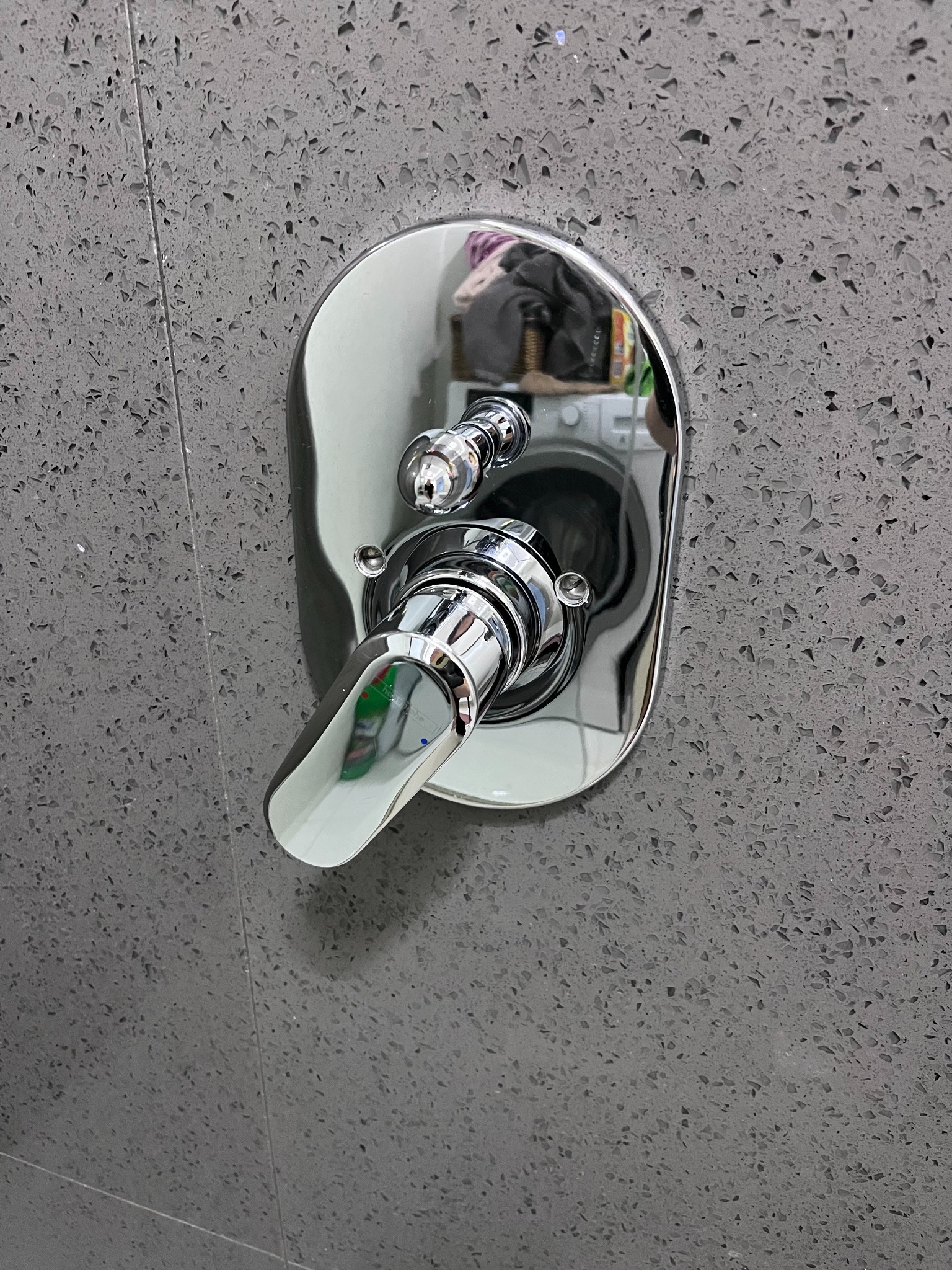 Bateria prysznicowa chromowana, zamontowana na szarej, teksturowanej ścianie. W odbiciu widać pralkę i inne przedmioty.