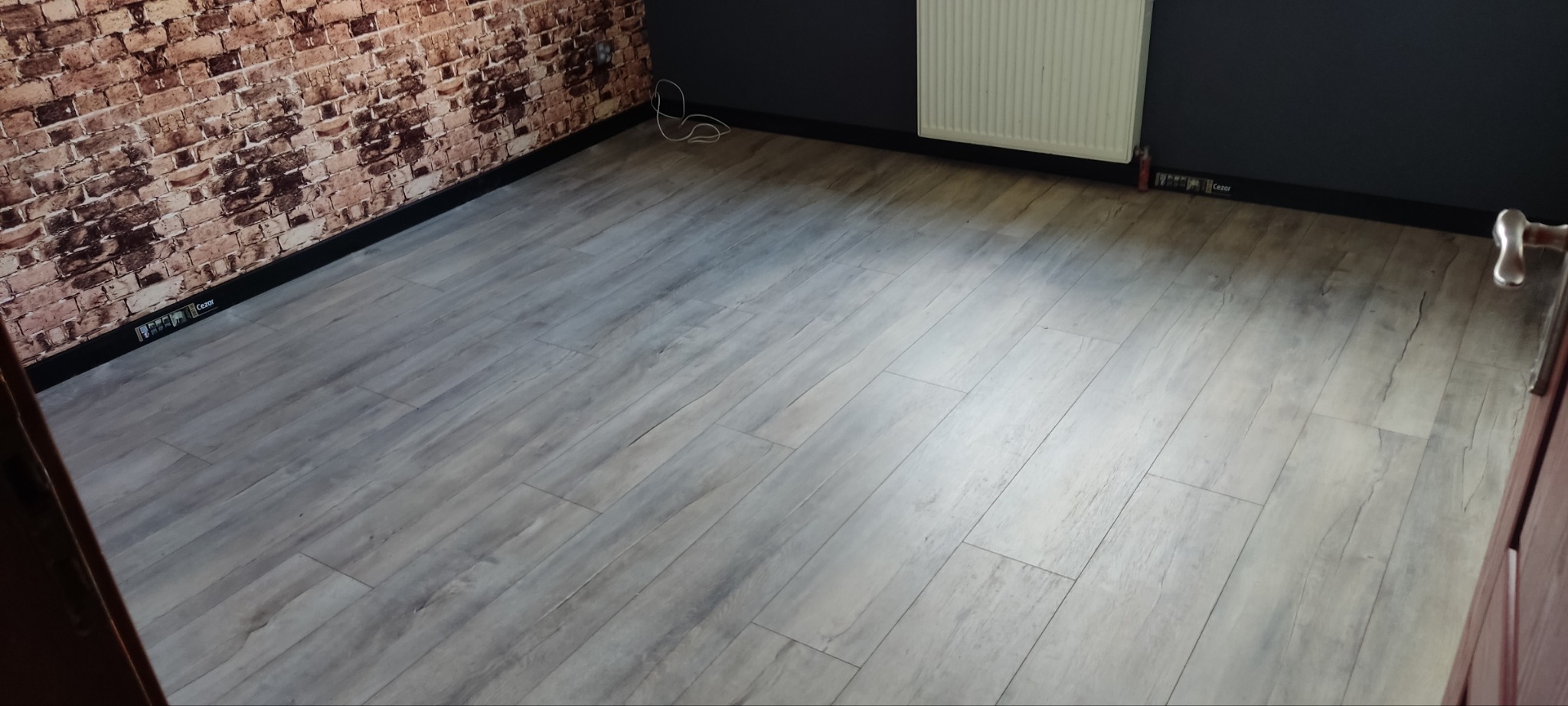 Jasnoszare panele podłogowe ułożone w pokoju z fragmentem ściany imitującej czerwoną cegłę i ciemnoszarą ścianą, widoczny grzejnik i listwy przypodłogowe.