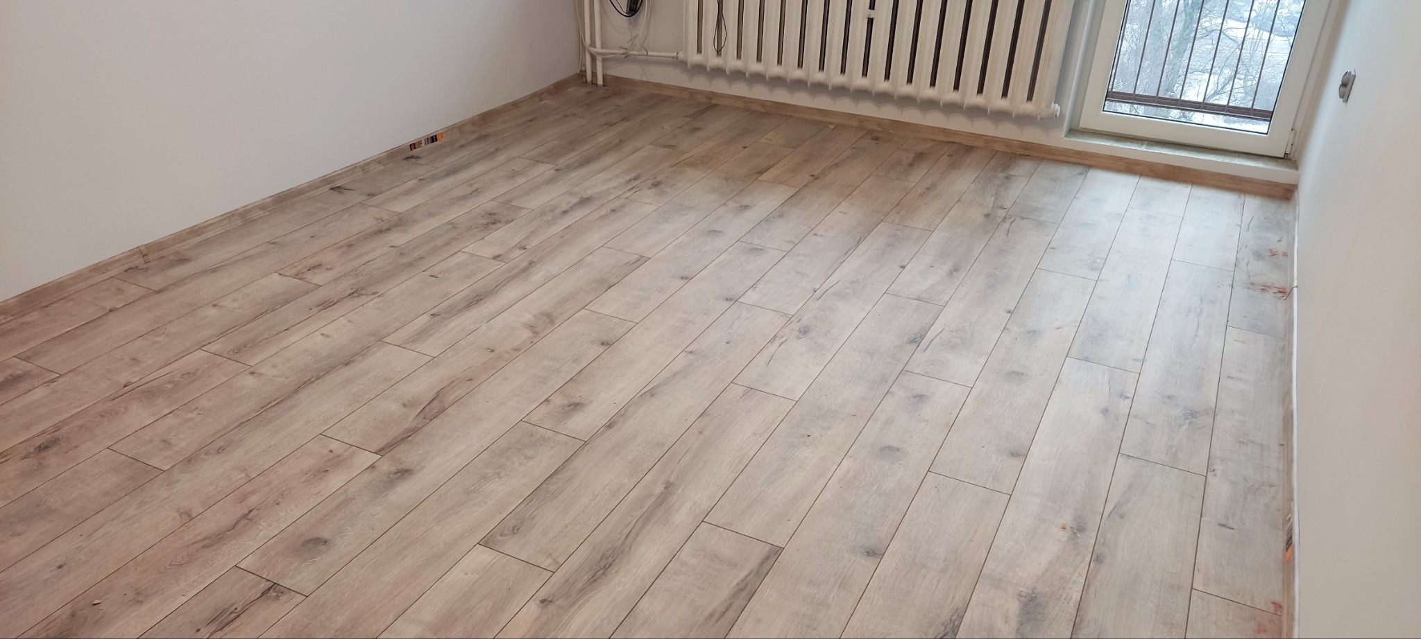 Jasne panele podłogowe imitujące drewno, ułożone w pokoju z grzejnikiem i oknem, widoczne drobne niedoskonałości przy ścianie.