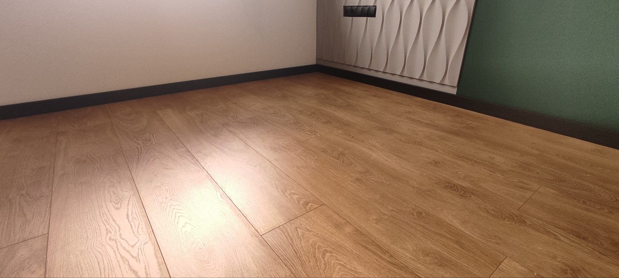 Nowo ułożona podłoga z paneli w odcieniach brązu, z widocznymi listwami przypodłogowymi w kolorze czarnym, ściana w kolorze kremowym oraz dekoracyjna ściana 3D.