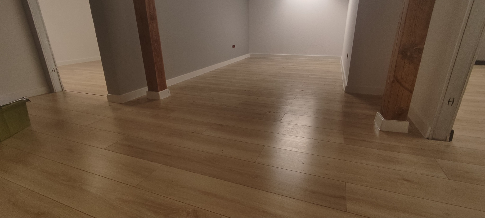 Jasne panele podłogowe ułożone w pustym pomieszczeniu z drewnianymi słupami i białymi listwami przypodłogowymi.