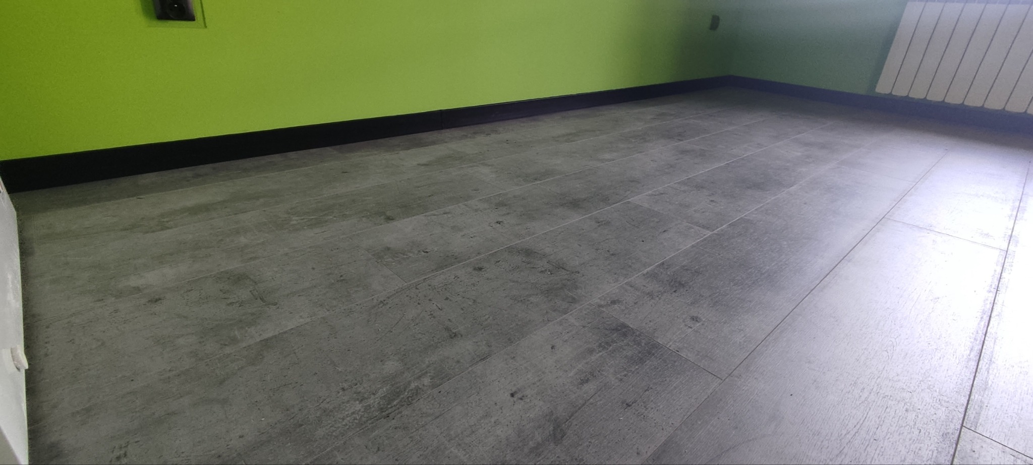 Szare panele podłogowe ułożone w pomieszczeniu z limonkową ścianą i czarną listwą przypodłogową, widoczny fragment białego grzejnika.