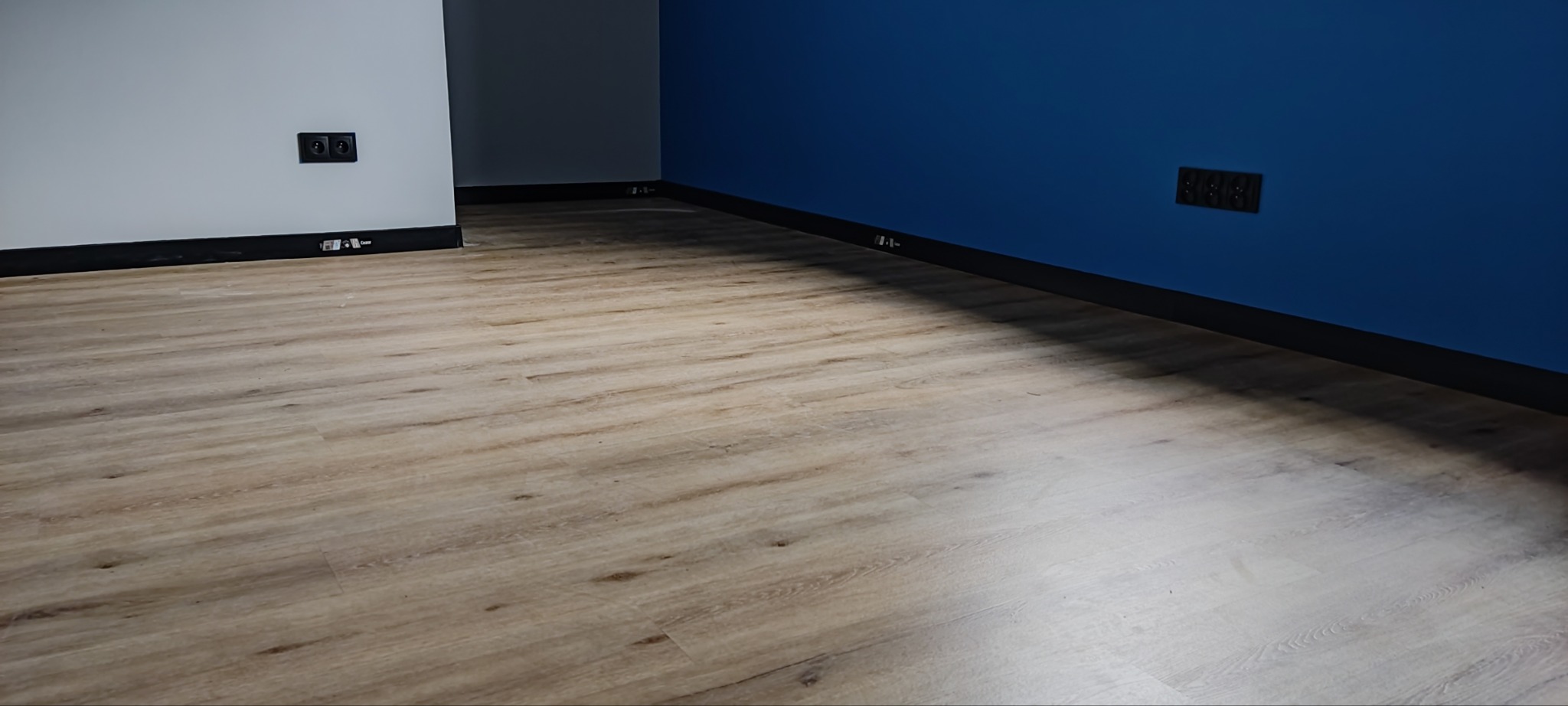 Ułożone panele podłogowe w jasnym odcieniu drewna, widoczne listwy przypodłogowe w kolorze czarnym oraz gniazdka elektryczne na ścianach w kolorach szarym i granatowym.