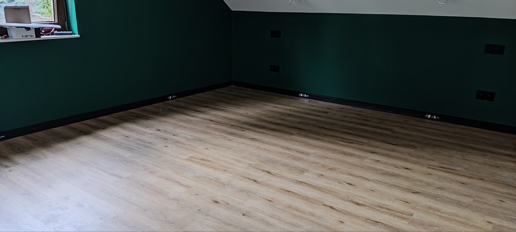 Ukończone wnętrze: jasne panele podłogowe ułożone w pokoju z ciemnozielonymi ścianami i czarnymi listwami przypodłogowymi oraz gniazdkami elektrycznymi.
