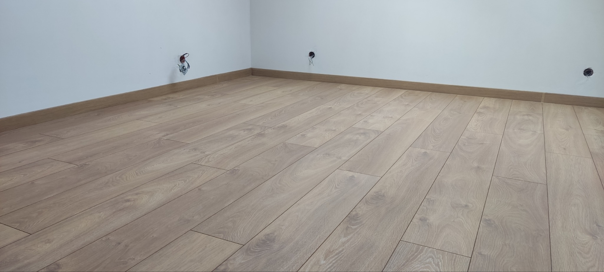 Jasne panele podłogowe ułożone w pustym pomieszczeniu, widoczne listwy przypodłogowe i wystające przewody elektryczne ze ściany.