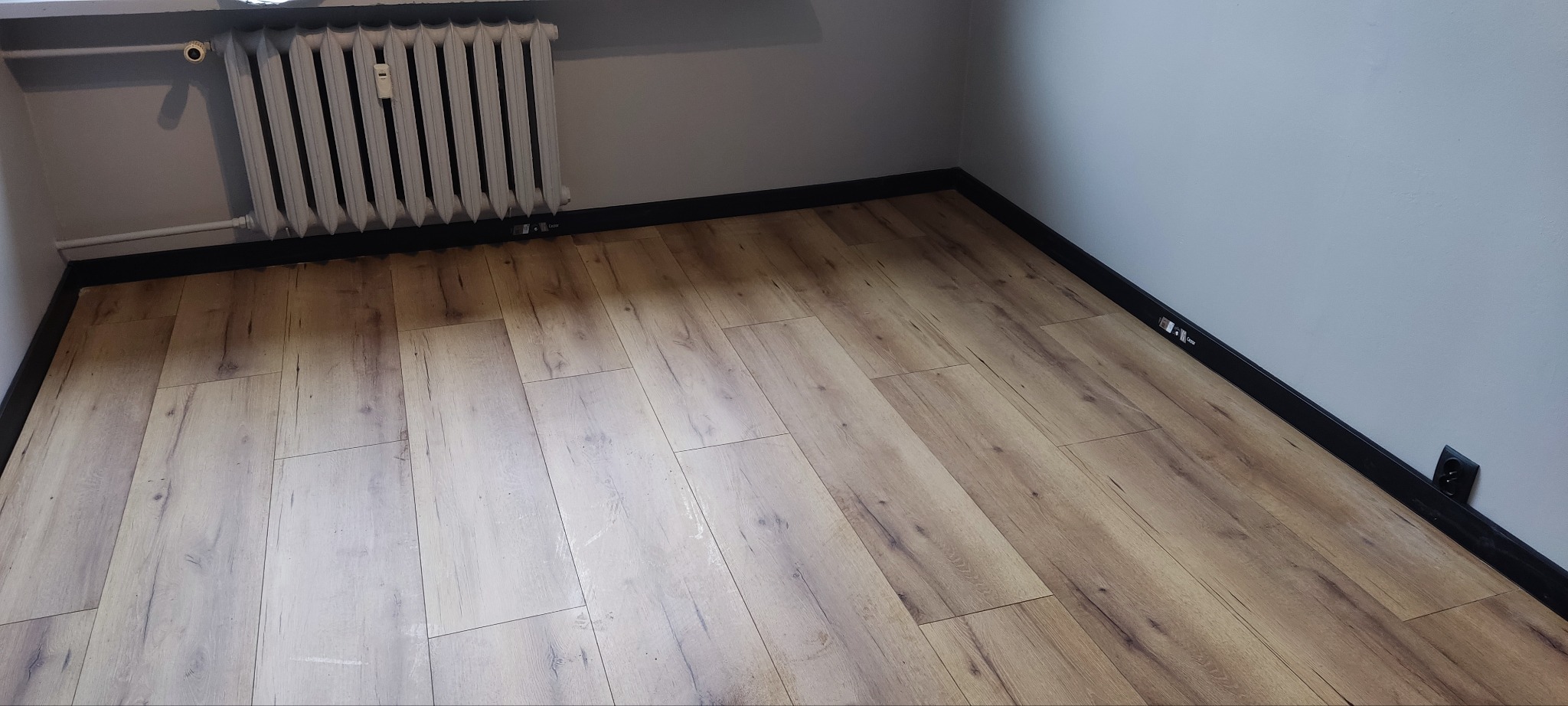 Ułożone panele podłogowe w rogu pokoju, widoczny grzejnik i czarne listwy przypodłogowe.