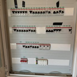 Instalatorstwo Elektryczne Kacper Szymala - Otwarta skrzynka rozdzielcza z bezpiecznikami, oznaczone numery od 1 do 50, niektóre zabezpieczone czarną taśmą, widoczne różne typy wyłączników, na dole fragment drewnianej ściany i niebieskiego...