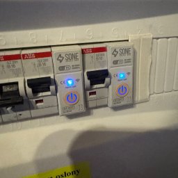Instalatorstwo Elektryczne Kacper Szymala - Szczegółowe ujęcie tablicy rozdzielczej z zamontowanymi modułowymi wyłącznikami nadprądowymi SONE Smart One z włączonymi diodami LED, widoczna żółta naklejka z napisem.