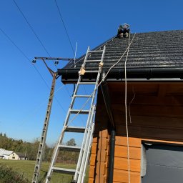 Instalatorstwo Elektryczne Kacper Szymala - Pracownik w kasku klęczy na dachu drewnianego domu, obok drabina oparta o rynnę, widoczny słup energetyczny i niebieskie niebo.